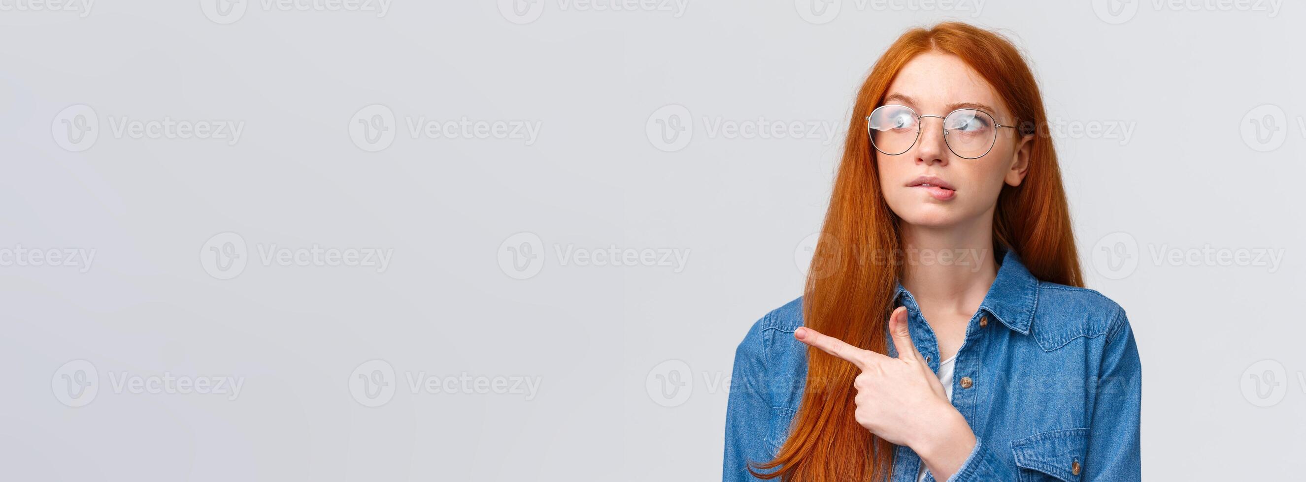 Unsure smart and cute redhead teenager in glasses, female student biting lip curious looking and pointing left, thinking, deciding what pick, making choice, standing over white background photo