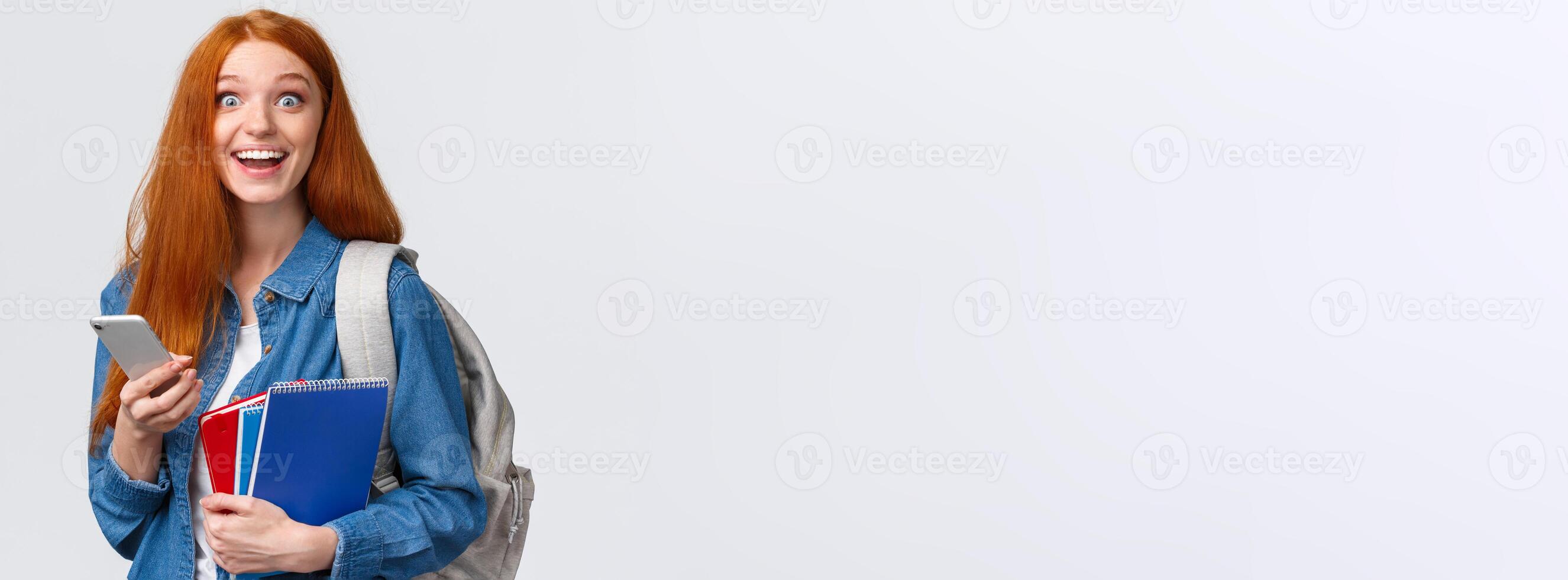 Excited and happy, relieved cute redhead female college student with backpack, notes in notebook and smartphone, looking cheerful camera, texting, messaging friends, white background photo