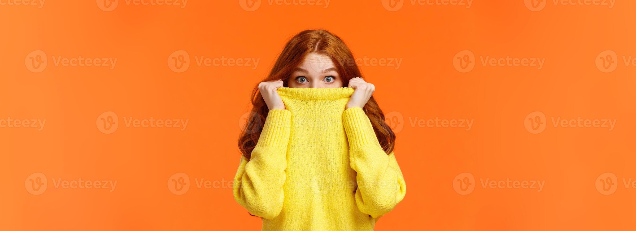 Surprised and astonished cute curious redhead female pulling sweater collar on face and popping eyes at camera with amazed expression, found something interesting, orange background photo