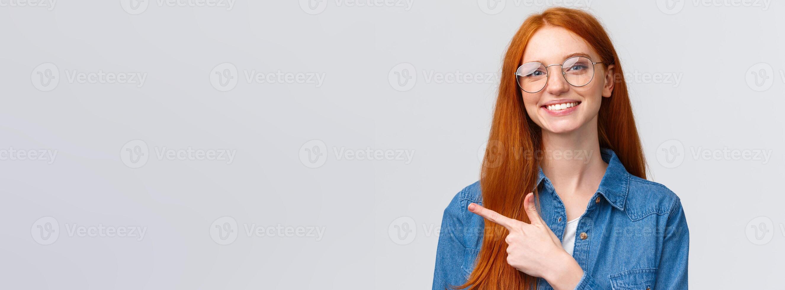 Close-up portrait cute cheerful redhead, foxy girl in glasses, showing place to click website, follow or subscribe, pointing finger left and smiling pleasant, with friendly attitude, white background photo