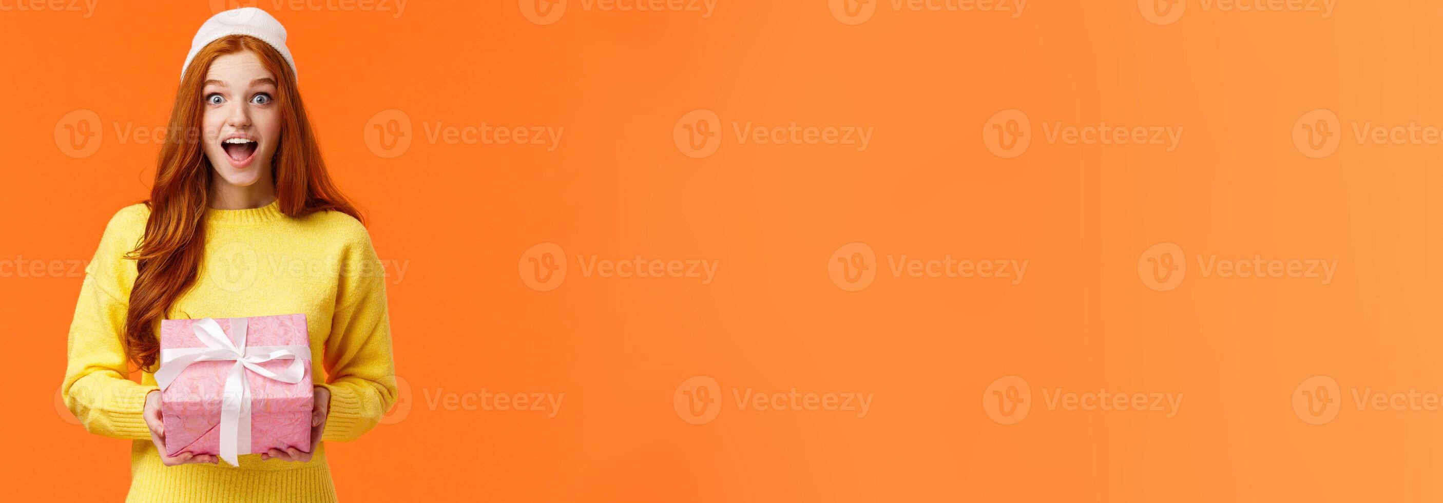 Surprised and excited happy girl unwrapping gifts on christmas eve, new year celebration, smiling amused and thrilled, holding wrapped box present, wearing winter beanie, orange background photo