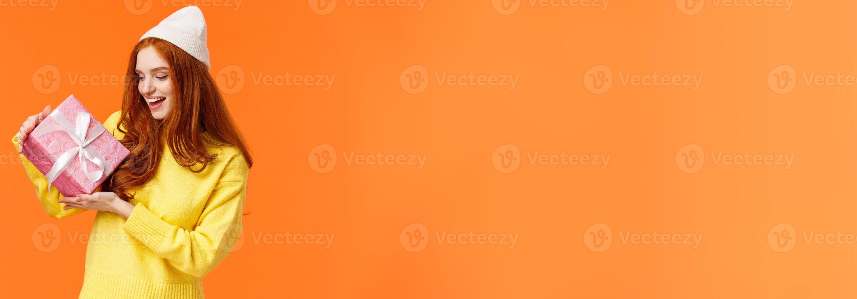 Girl trying figure-out whats inside gift box shaking it and looking intrigued. Excited happy redhead woman like winter holidays and receiving present, standing amused orange background photo