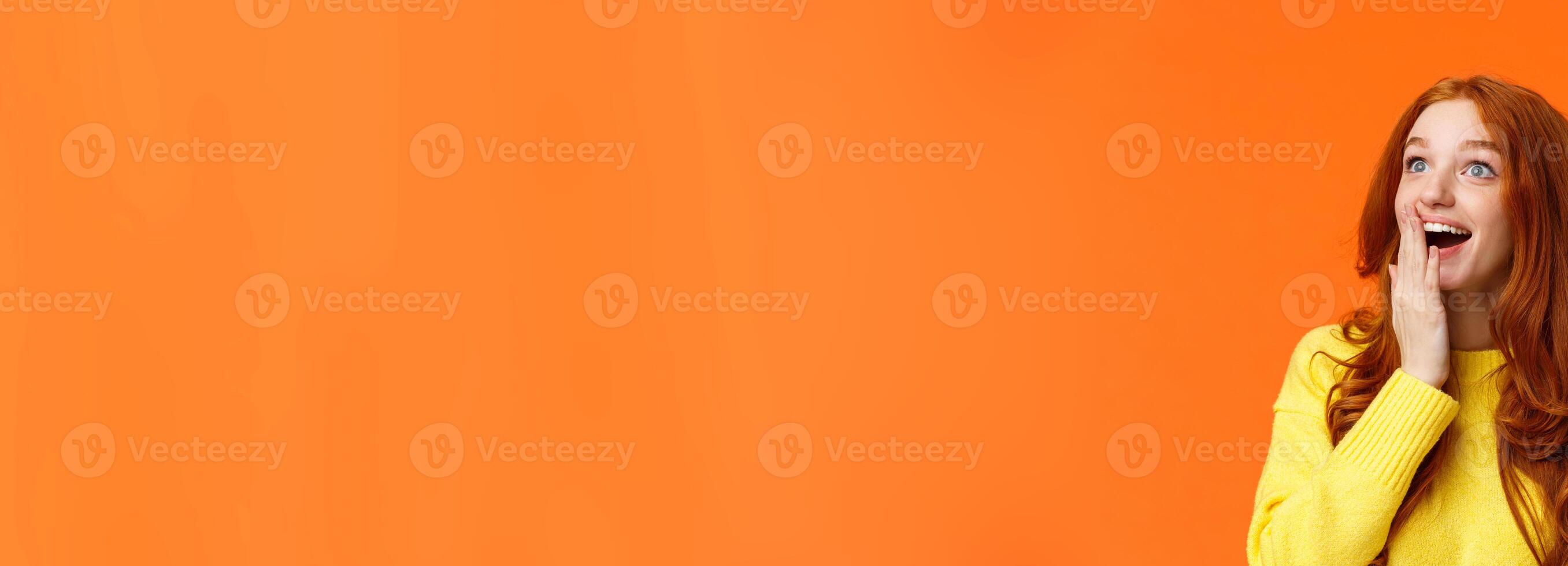 Fascinated and amiring cute redhead woman looking impressed and excited at upper left corner, smiling see breathtaking view, cover opened mouth palm thrilled, losing speech happy, orange background photo