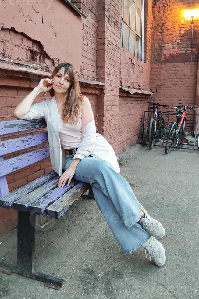a sweet, calm woman with dark, dyed hair, in a white jacket, sits on a bench in the city, waiting for a meeting photo