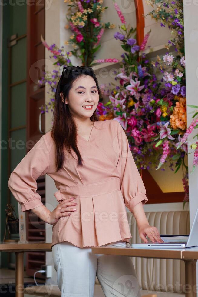 Charm of beautiful young indonesia woman cheerful holding waist and laptop looking at camera standing in front of sofa in indoor coffee shop. for fashion, lifestyle, promotion, advertising content photo