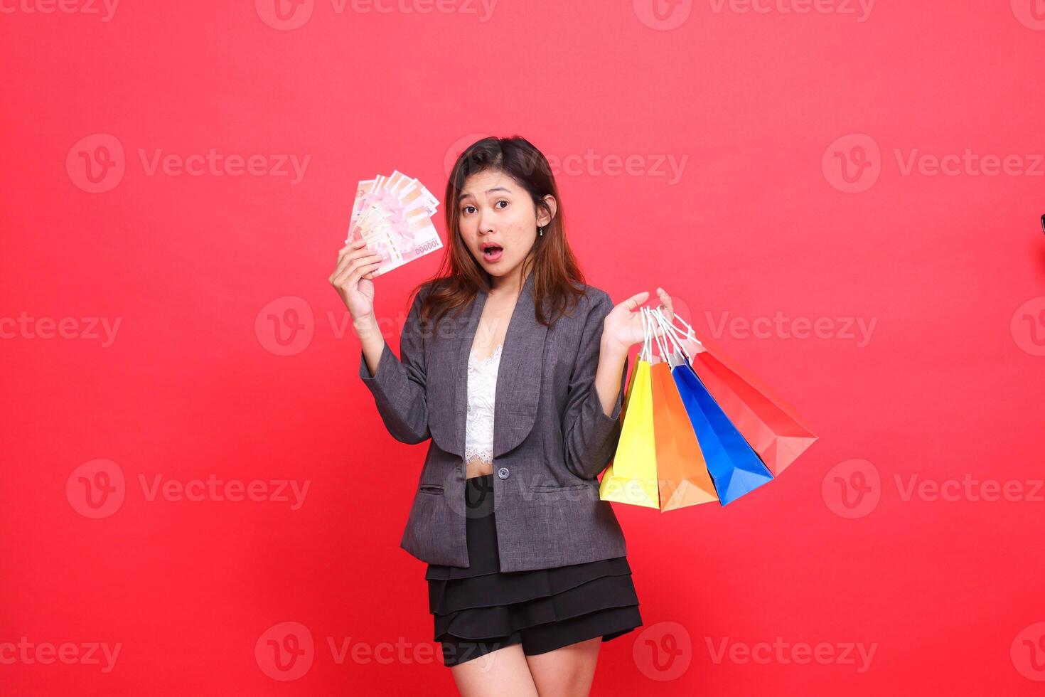 Asian office woman shocked at the camera carrying rupiah money and holding various shopping paper bags wearing a gray jacket with a red background. for transaction, lifestyle and business concepts photo