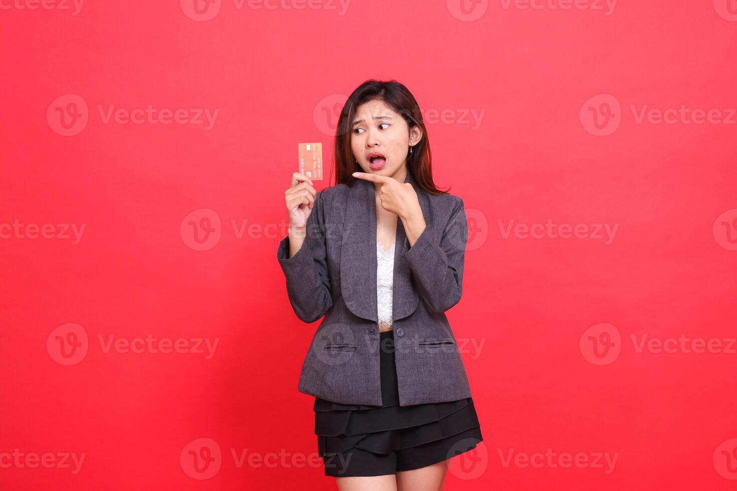Asian office woman's expression shocked candid hand holding a debit credit card while pointing at it wearing a jacket and skirt on a red background. for financial, business and advertising concepts photo