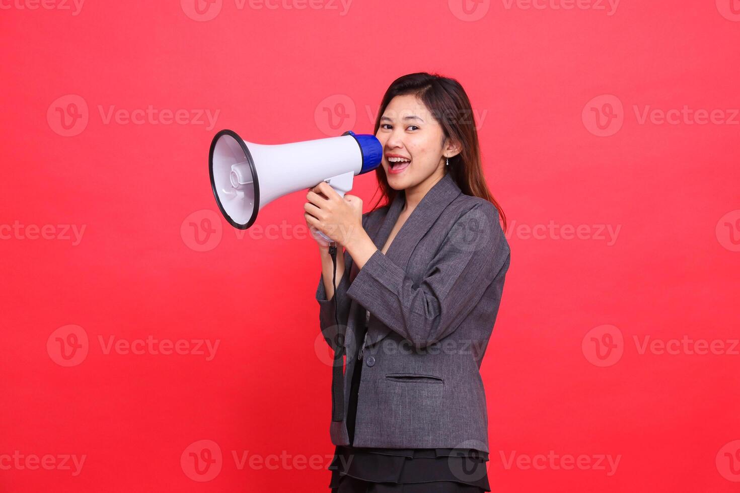 The expression of a cheerful Asian office woman leaning to the right shouts using a megaphone into the camera wearing a gray jacket and skirt on a red background. for broadcast and advertising photo