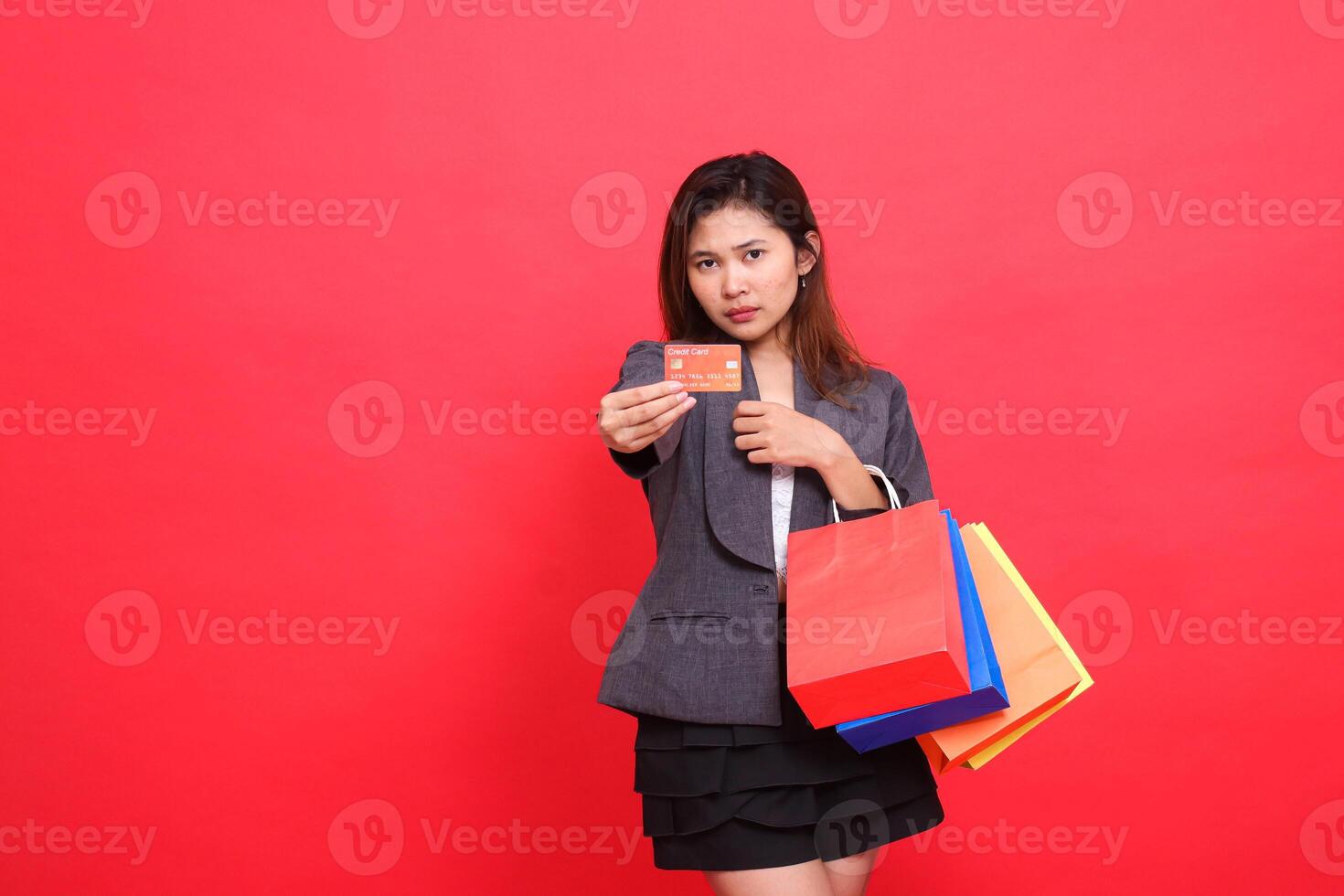sad asian office woman carrying shopping paper bag while holding credit debit card forward wearing formal jacket with red background. for transaction, lifestyle and business concepts photo