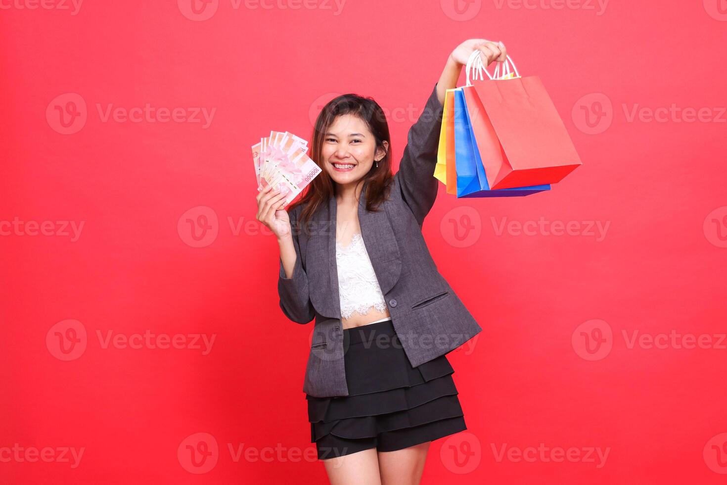 Cheerful Asian office woman looking at the camera carrying rupiah money and holding up various shopping paper bags wearing a gray suit on a red background. for transaction, lifestyle and business photo