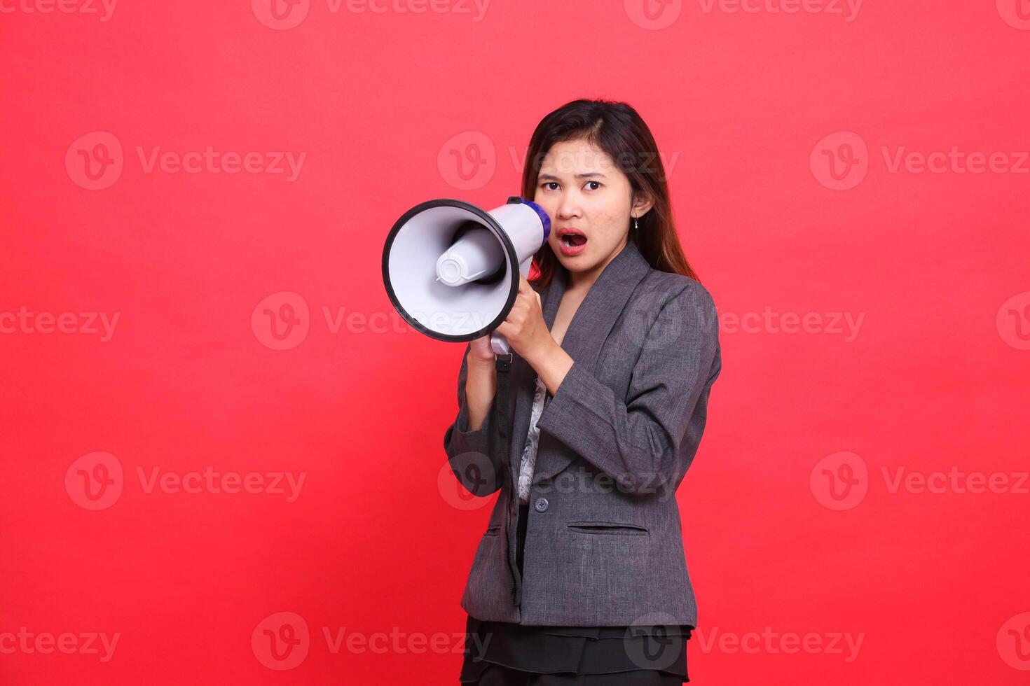 Asian office girl expression is very angry screaming using a megaphone in front of her wearing a gray jacket and skirt on a red background. for broadcast, business and advertising concepts photo