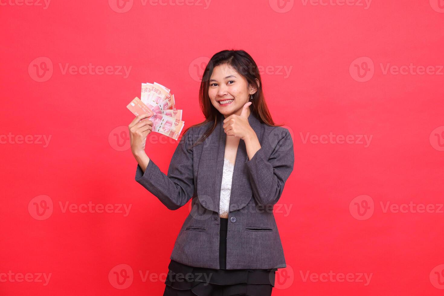 indonesia office woman smiling, holding credit, debit card and money while giving a thumbs up sign to the camera wearing a gray jacket and red skirt. for transaction, business and advertising concepts photo