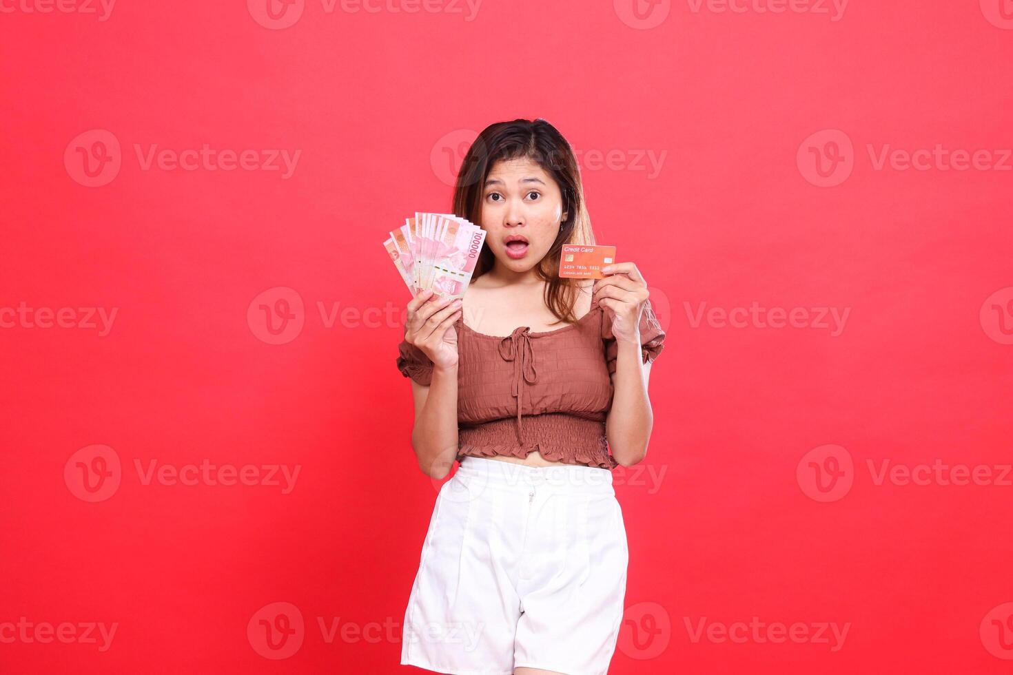 Shocked indonesia woman holding rupiah money and debit credit card wearing brown blouse top with red background. for transaction, technology and advertising concepts photo