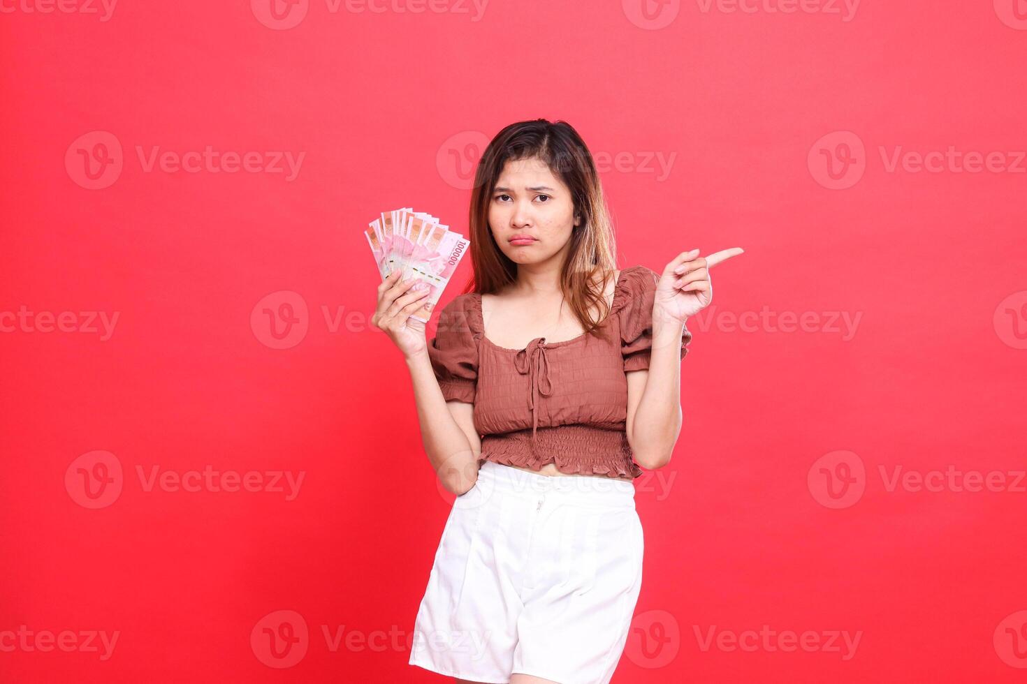 The expression of a sullen Asian girl holding rupiah money while pointing to the left wearing a brown blouse with a red background. for transaction, technology and advertising concepts photo