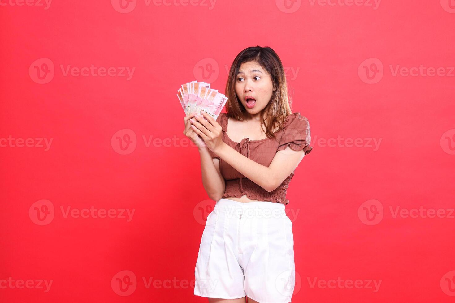 Asian woman's expression, shocked, happy, candid, both hands holding rupiah currency, wearing a brown blouse, red background. for transaction, technology and advertising concepts photo