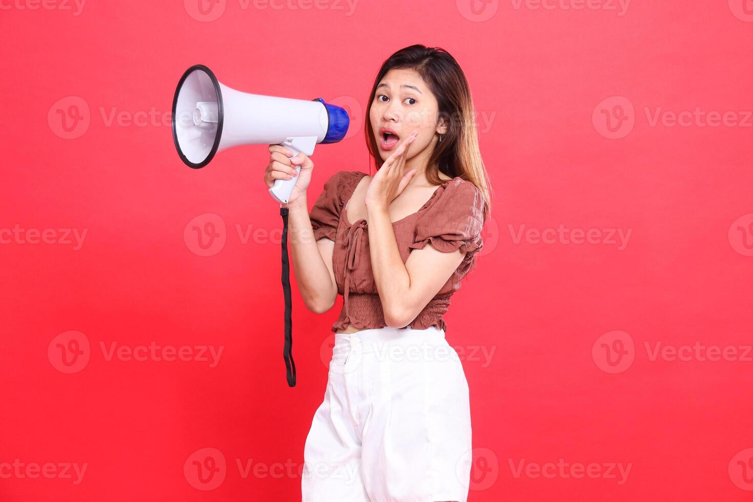 the expression of an indonesia woman screaming in surprise at the camera holding an audio megaphone while focusing on the sound wearing a brown blouse top and shorts on a red background technology photo