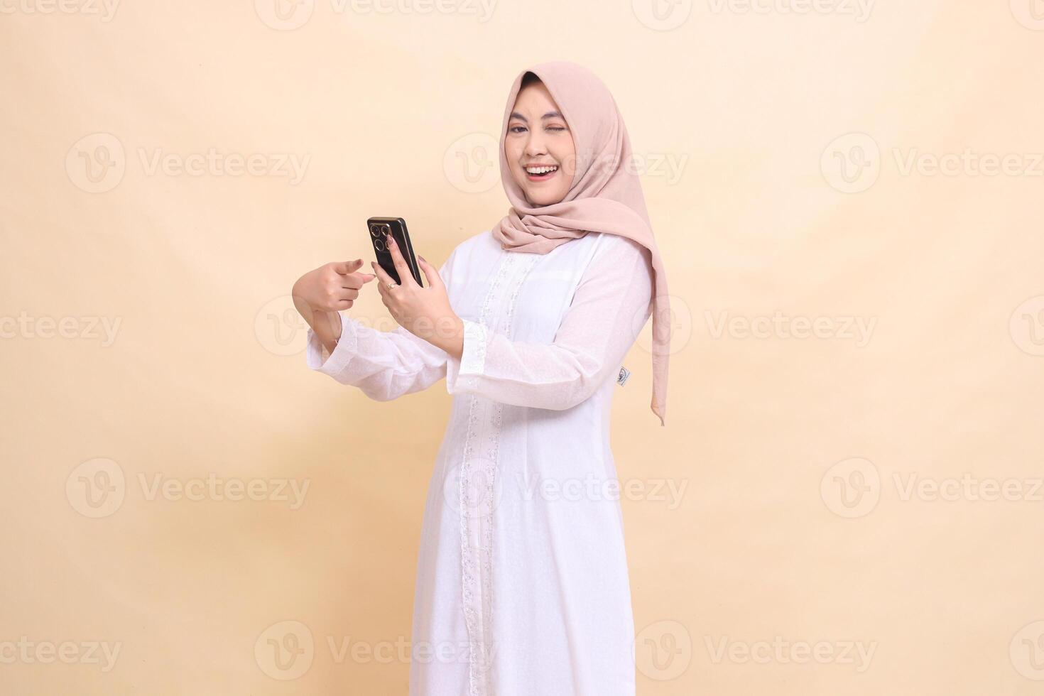 Asian Muslim woman wearing a hijab smiling happily winking at the camera holding a cell phone gadget in her hand while pointing at it. Lifestyle, technology and promotion concept photo