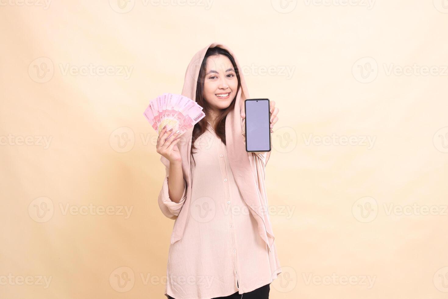 indonesia woman smiling cheerfully at the camera carrying rupiah money and holding the cellphone gadget screen in front of her. beautiful young for finance, technology, fashion photo