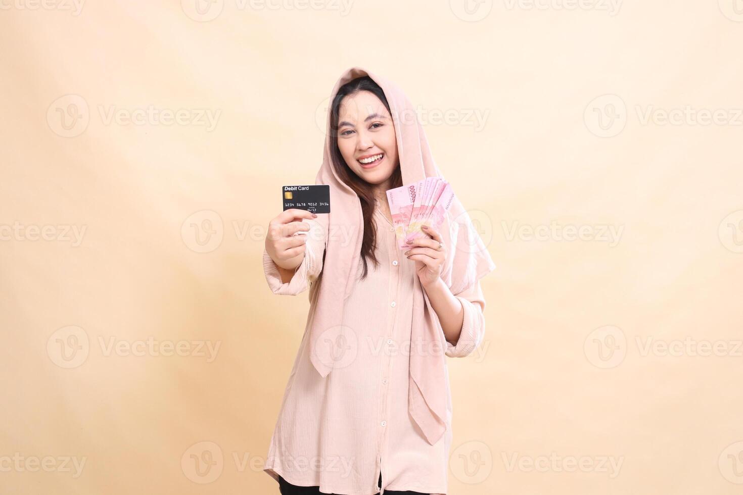 Asian woman with smiling expression laughing at you, holding your debit card in front of you and carrying rupiah money. young beautiful for finance, advertising, fashion photo