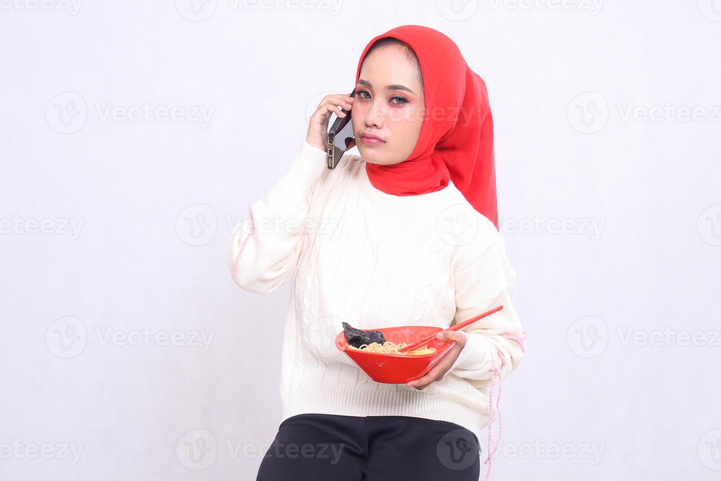 disappointed Asian woman wearing a hijab calls with a cellphone and carries a bowl containing ramen Chinese food and chopsticks. Beautiful Muslim women are used for culinary and advertising content photo