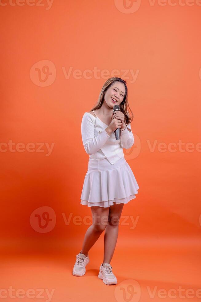 full length beautiful indonesia woman white top standing singing cheerfully to camera both hands holding mic audio isolation orange background. for fashion, advertising, lifestyle, entertainment photo