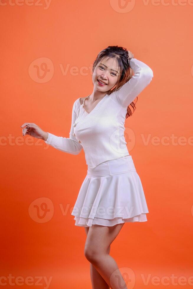 Beautiful Asian female model in white top standing cheerful wide slanted right hold hair back and dance pose isolated orange background. for fashion concept, advertisement, promotion, commercial photo