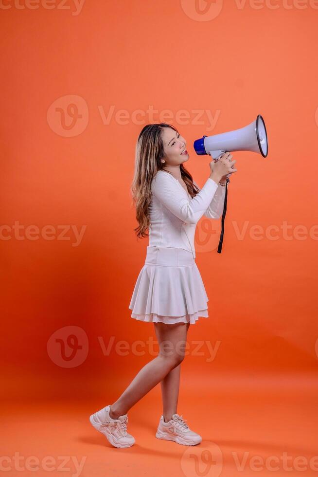 full length indonesia ladies white top standing left candid shouting cheerful both hands holding megaphone loudspeaker isolation orange background. for technology, advertising, broadcast photo