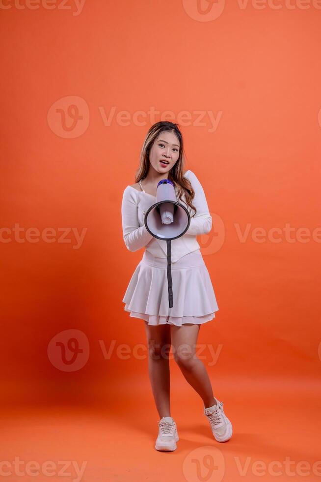 full length indonesia woman white top standing to camera screaming in surprise both hands holding megaphone loudspeaker diagonally down isolated orange background. for technology, advertising photo