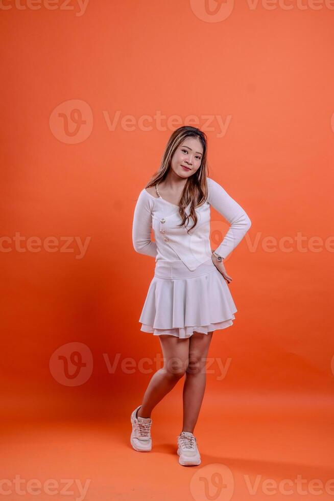full length beautiful asian woman white top standing smiling elegantly hold back and waist isolated orange background. for technology concept, advertising, broadcast, promotion photo