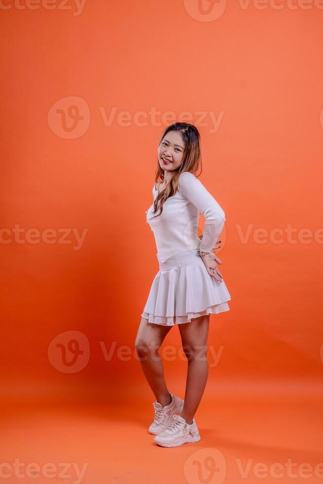full length beautiful indonesia woman white top standing smiling elegantly hold back and waist isolated orange background. for technology concept, advertising, broadcast, promotion photo
