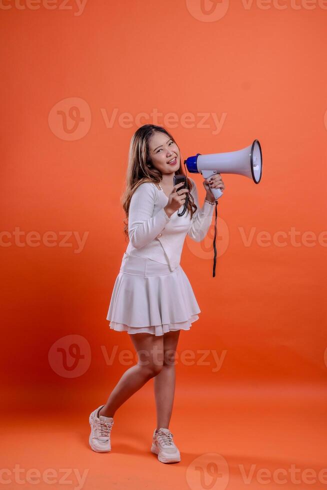 full body beautiful asian girl white top standing to camera screaming happily holding megaphone loudspeaker and mic audio isolation orange background. for technology, advertising, broadcast, promotion photo