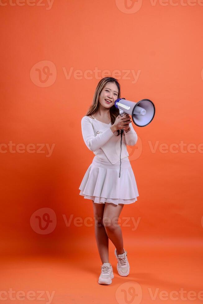 full length beautiful asian girl white top standing to camera screaming cheerful both hands holding megaphone loudspeaker isolated orange background. for technology, advertising, broadcast, promotion photo