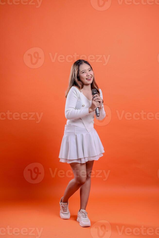 full body beautiful asian woman white top standing singing cheerfully in front of both hands holding mic isolated orange background. for fashion concept, advertising, lifestyle, entertainment photo