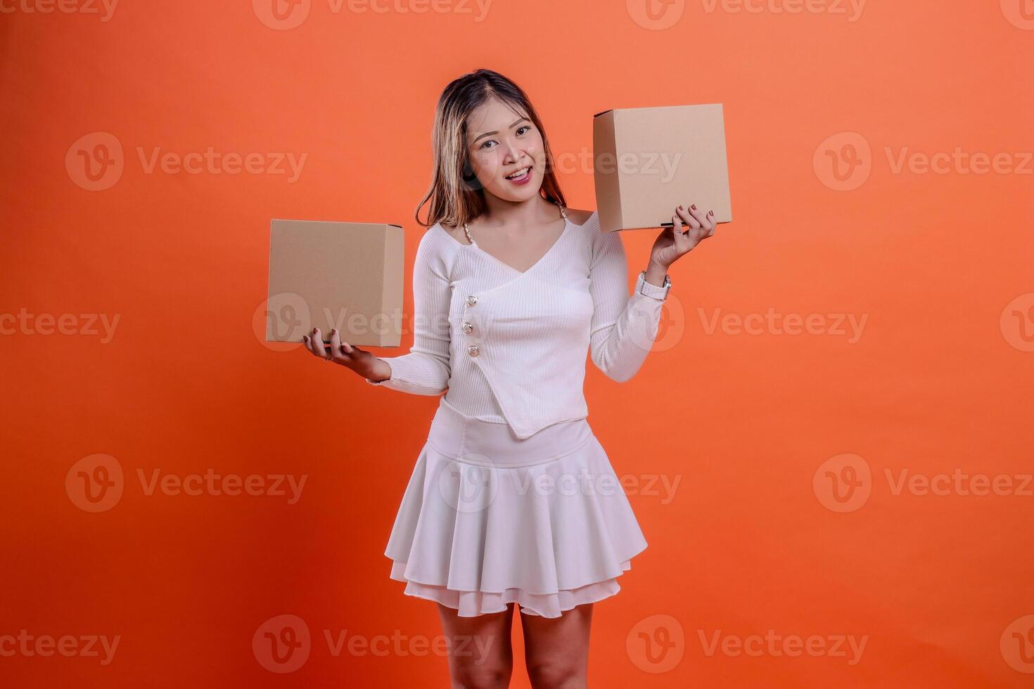 Beautiful adult female in white top happy to camera both hands lifting plain cardboard box up and down, standing on orange background. for delivery concept, gift, expedition, service, package photo