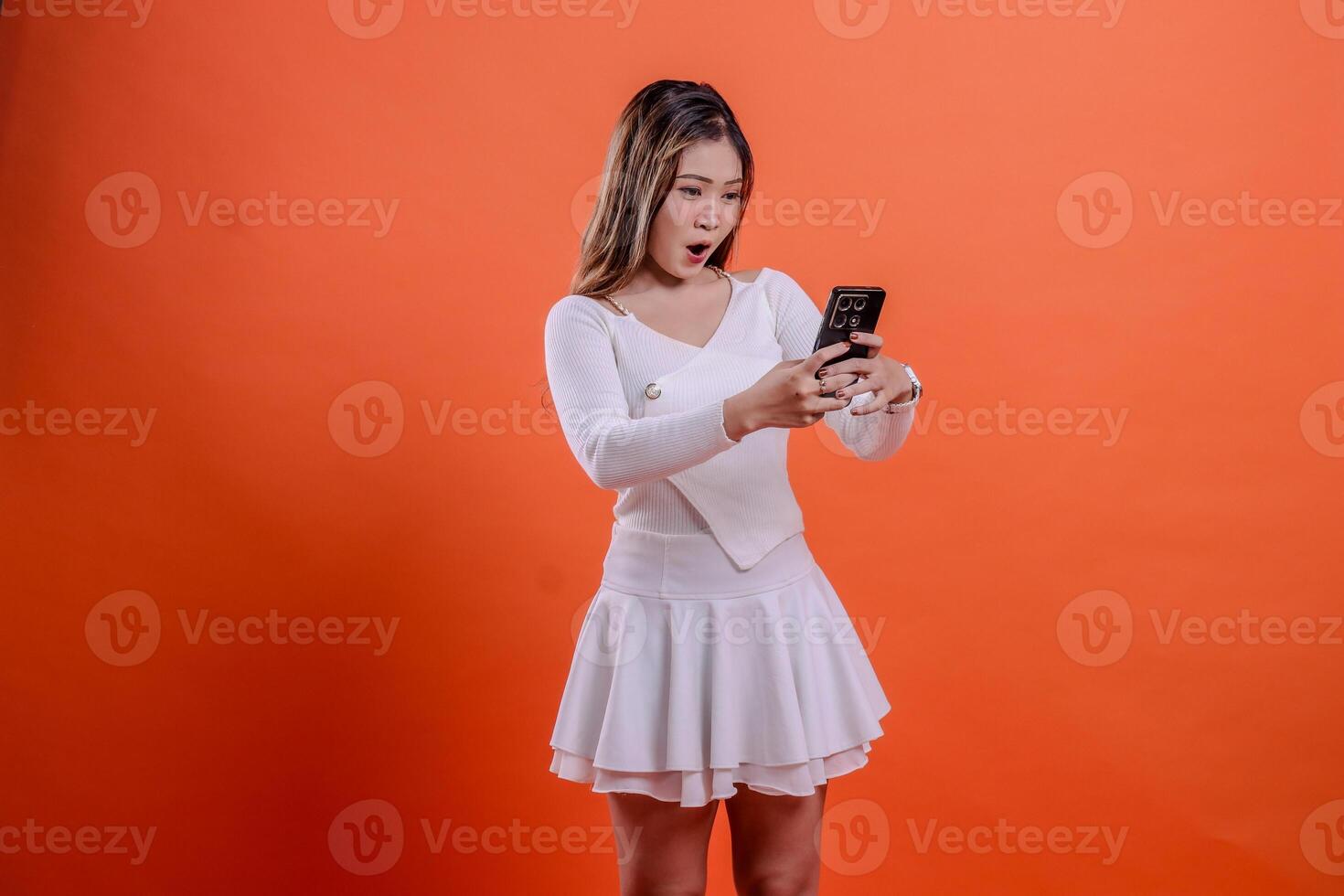 gesture beautiful young asian woman white top standing surprised excited to gadget slanted left both hands hold mobile phone, cellphone isolated orange background. for communication concept, business photo