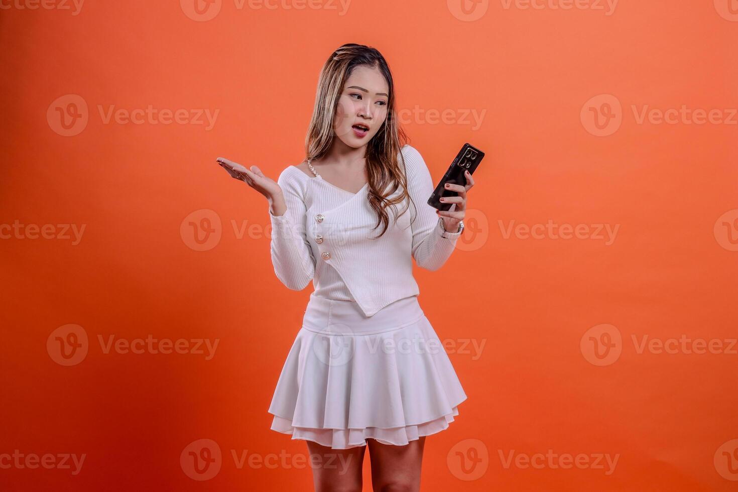 portrait of beautiful young asian woman in white top standing surprised to smart phone holding mobile phone, cellphone and hand sideways isolated orange background. for technology concept, transaction photo