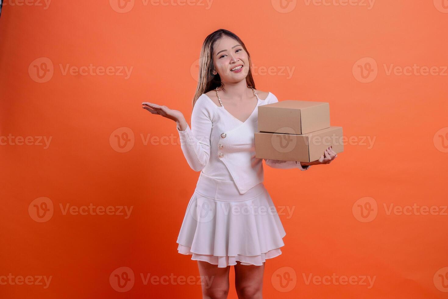 cheerful teenage woman expression in white top to camera hand carrying 2 plain cardboard boxes while presenting, standing on orange background. for delivery concept, gift, expedition, service, package photo