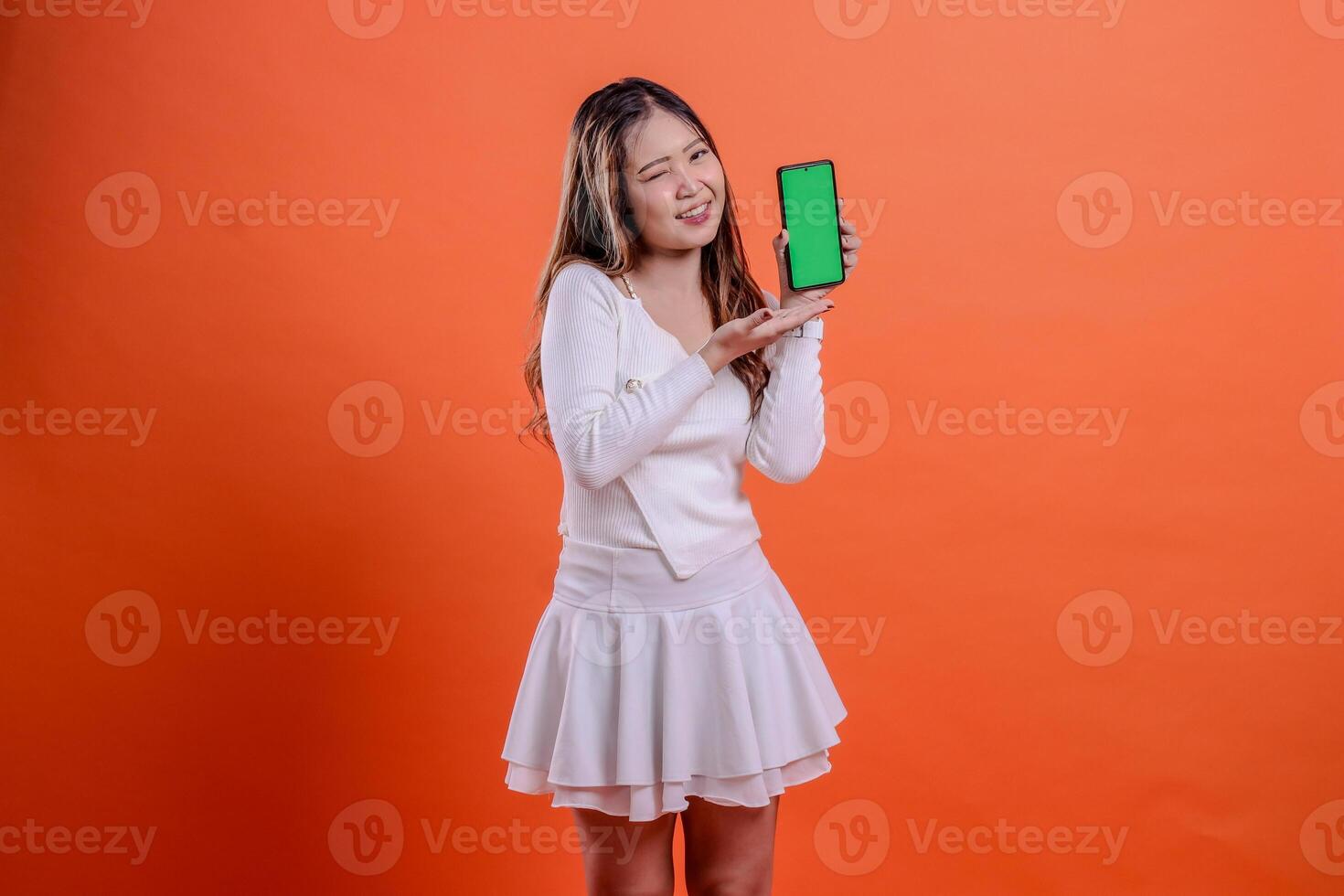 expression of asian woman in white top standing cheerful winking to camera hand holding mobile phone screen, cellphone and presenting isolated orange background. for technology, business, lifestyle photo