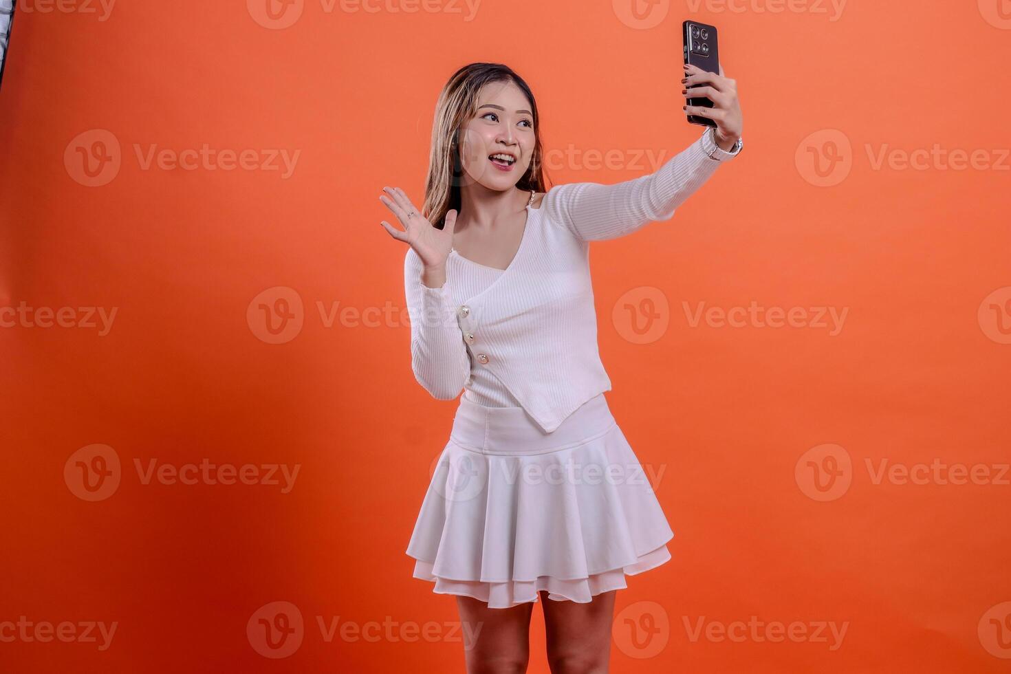 cheerful adult asian woman in white sweater call using smartphone camera, mobile phone, cellphone while pointing at face, standing. for technology, lifestyle, business, service photo