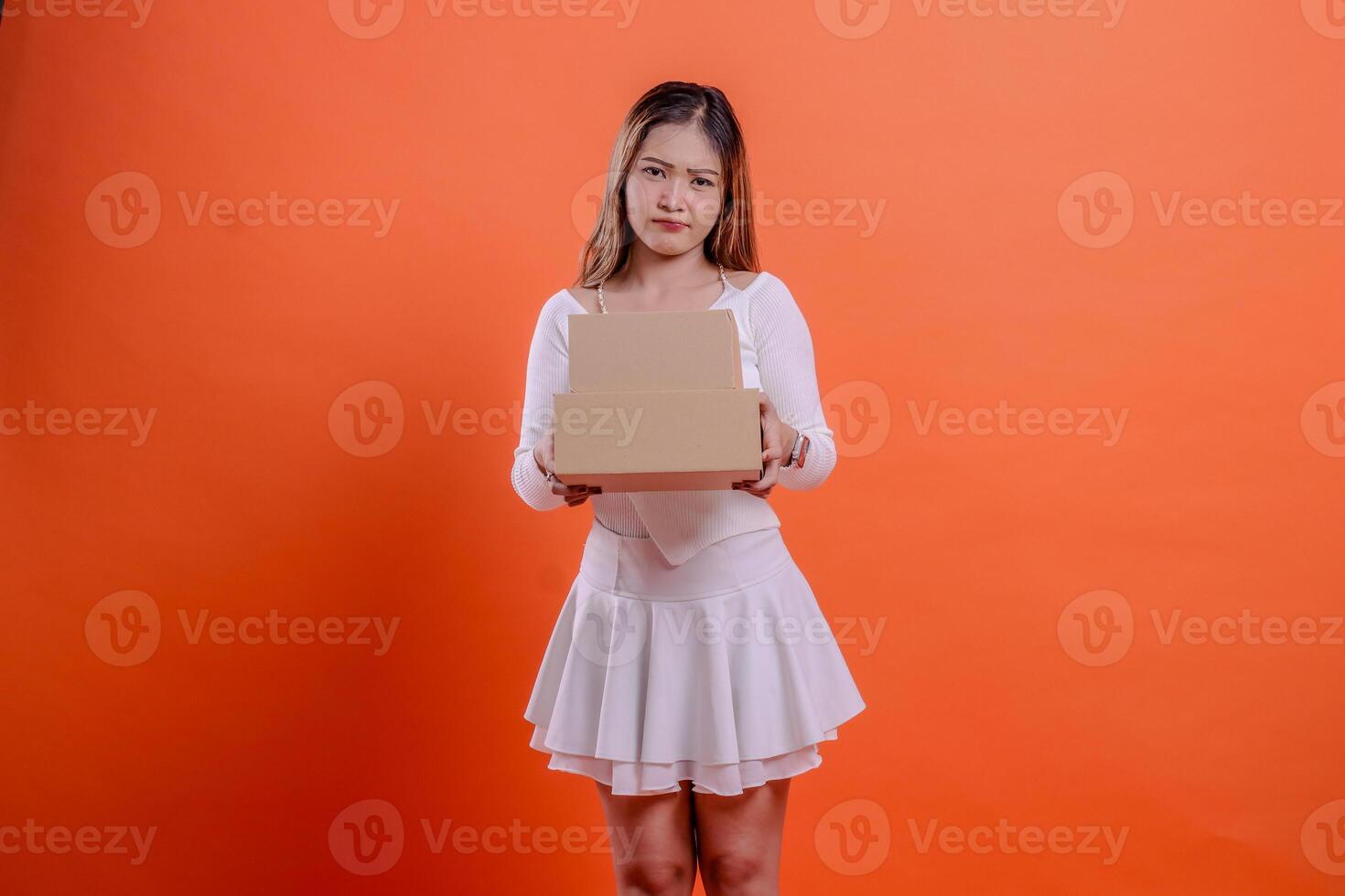 young asian girl in white top pouting at camera both hands holding 2 plain cardboard boxes, standing on orange background. for delivery, gift, expedition, service, package concept photo