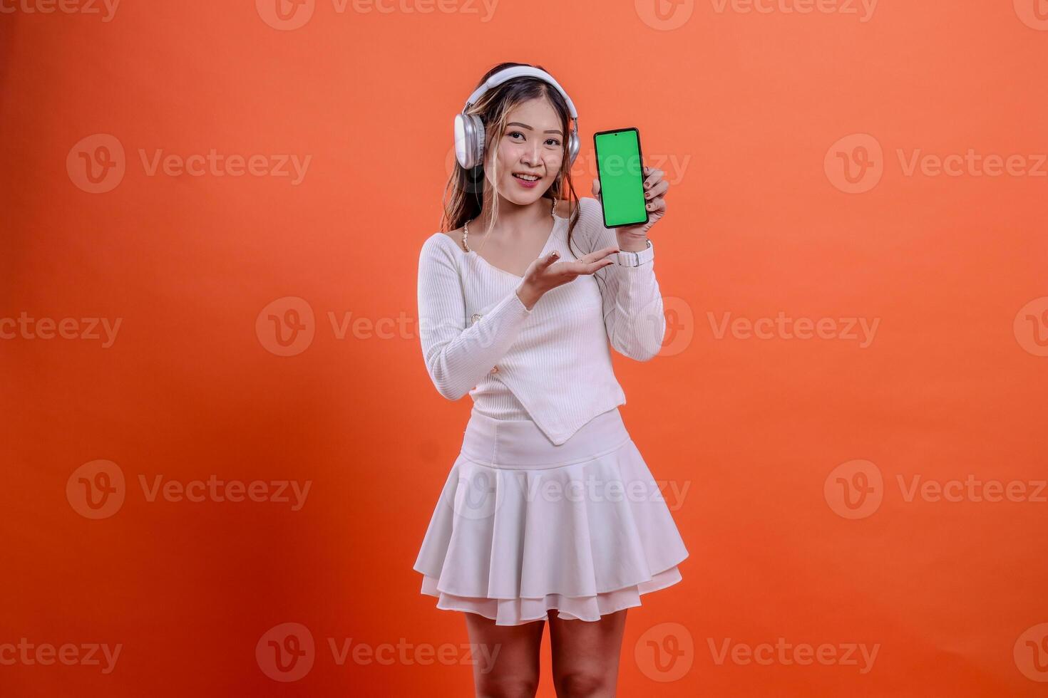 cheerful woman in white sweater wearing earphone headphone while holding mobile phone screen, cellphone green screen while presenting, standing on orange background. for entertainment, service concept photo