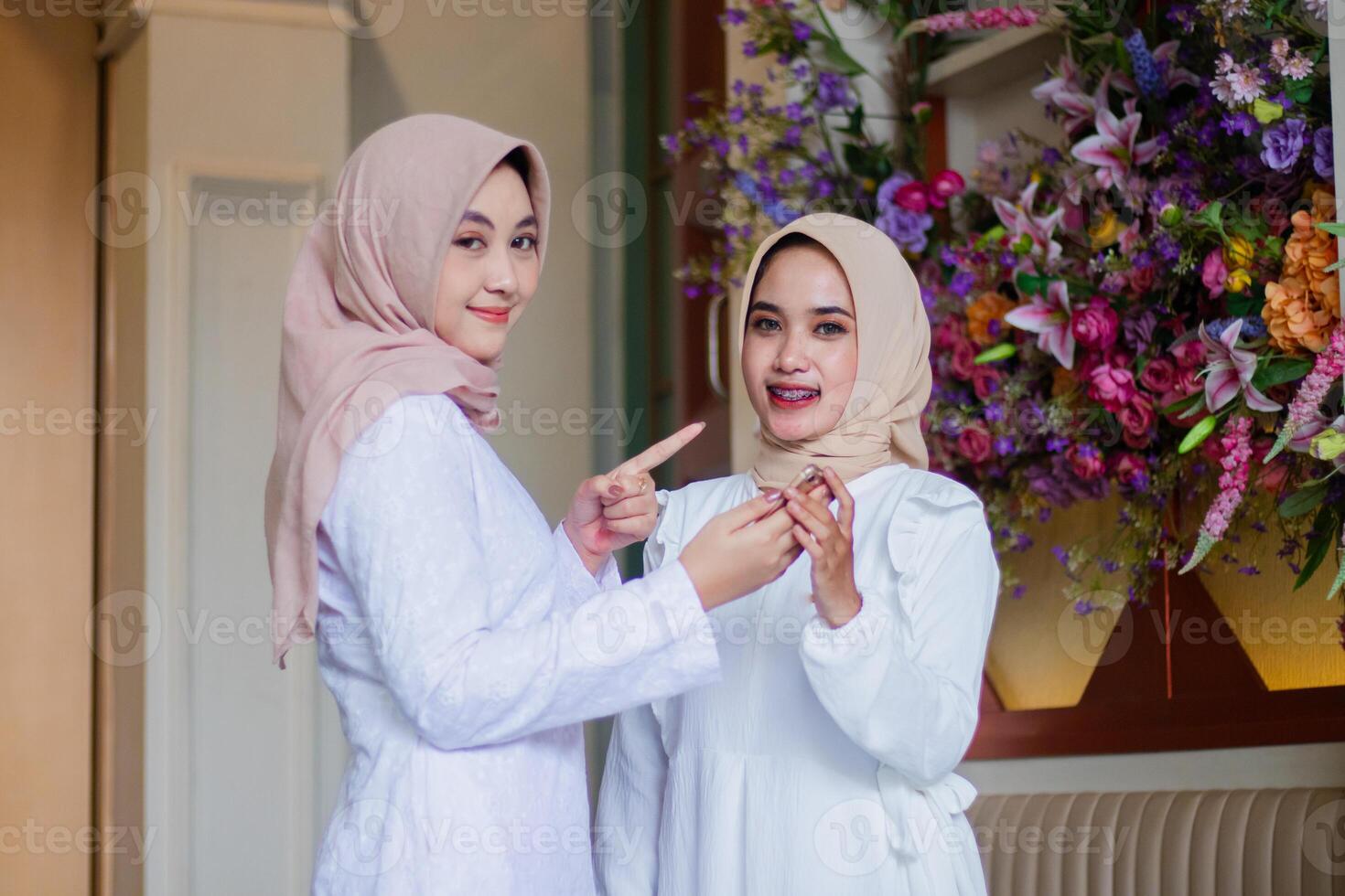 a group of young muslim girl wearing veil smile to the camera while holding mobile phone, cellphone, smart phone while pointing to the side standing in a restaurant. for technology content, lifestyle photo