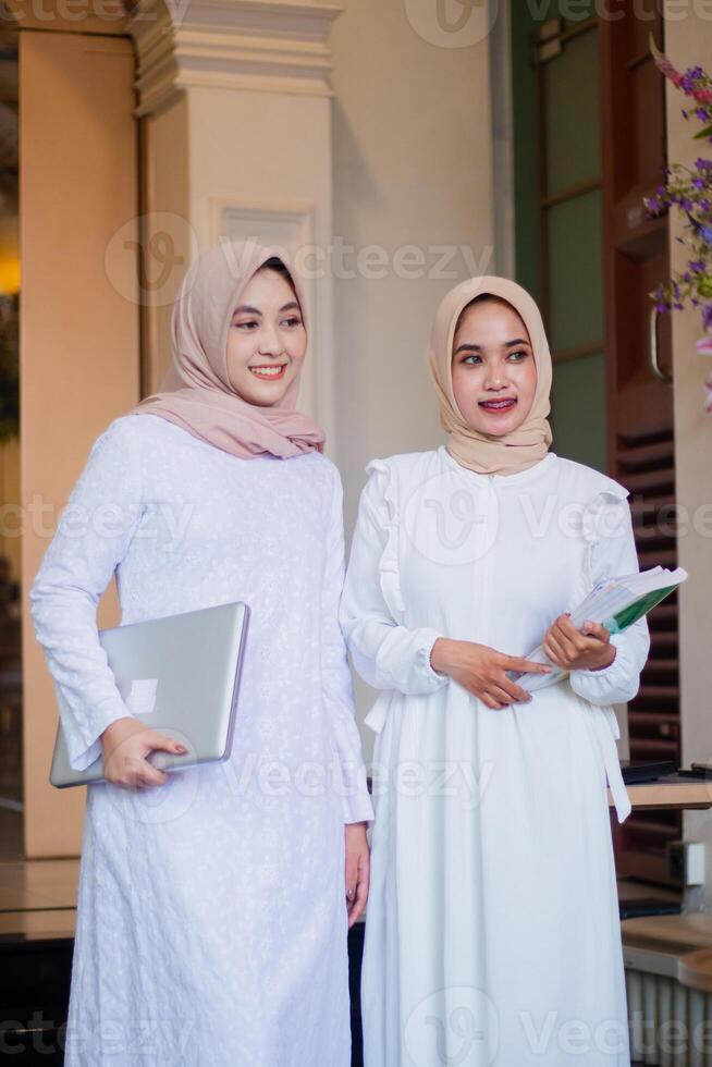 a pair of cheerful young Muslim girl in headscarf candid while holding a laptop and carrying a book beside standing in a coffee shop. for technology, education, lifestyle content photo