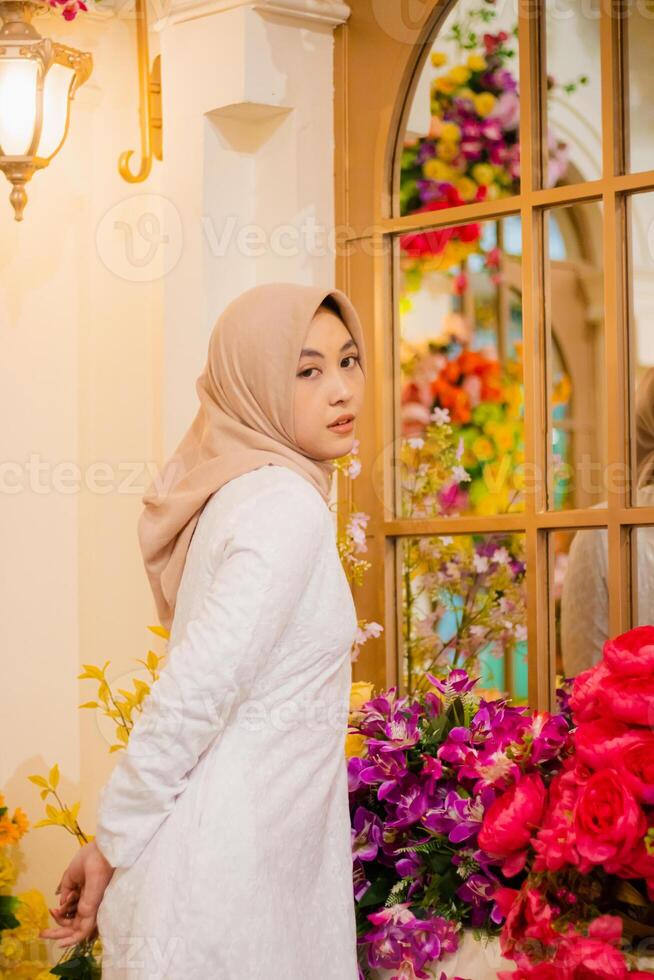 adult girl wearing hijab wearing a long white dress, elegant Muslim, to the camera, holding her chin and waist, standing among flower decorations in a luxury restaurant. for fashion, beauty, lifestyle photo