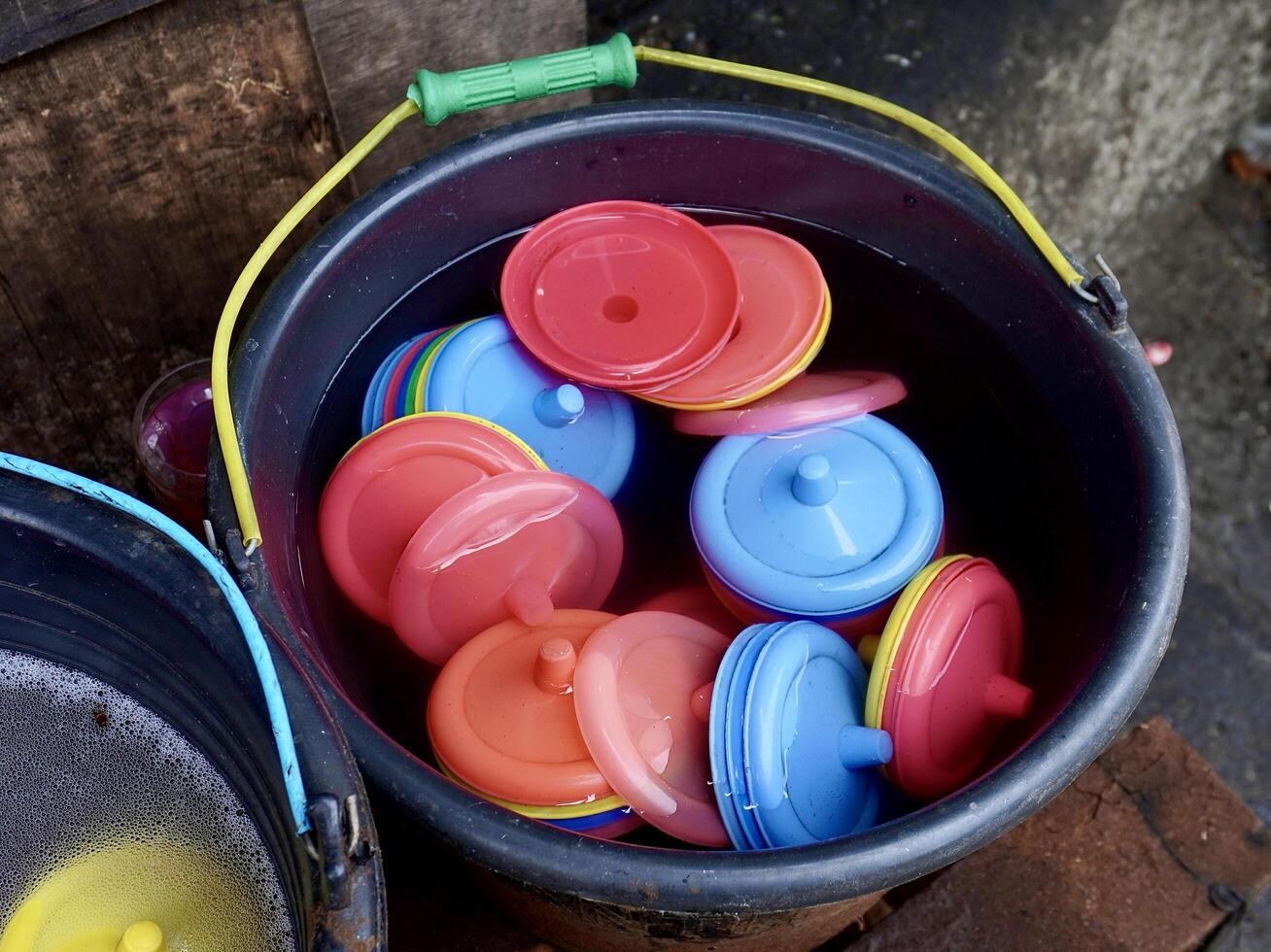 Colorful plastic glass lid or tutup gelas in black bucket filled with water isolated on horizontal ratio outdoor sidewalk dirty ground background. photo