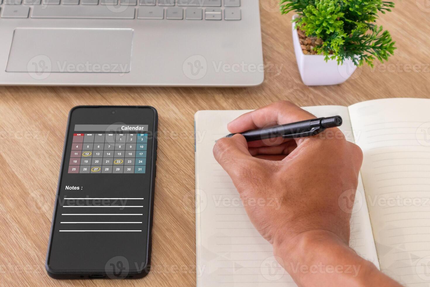 Man hand writing schedule calendar, reminder, or appointment in agenda book while looking at calendar app on mobile phone photo