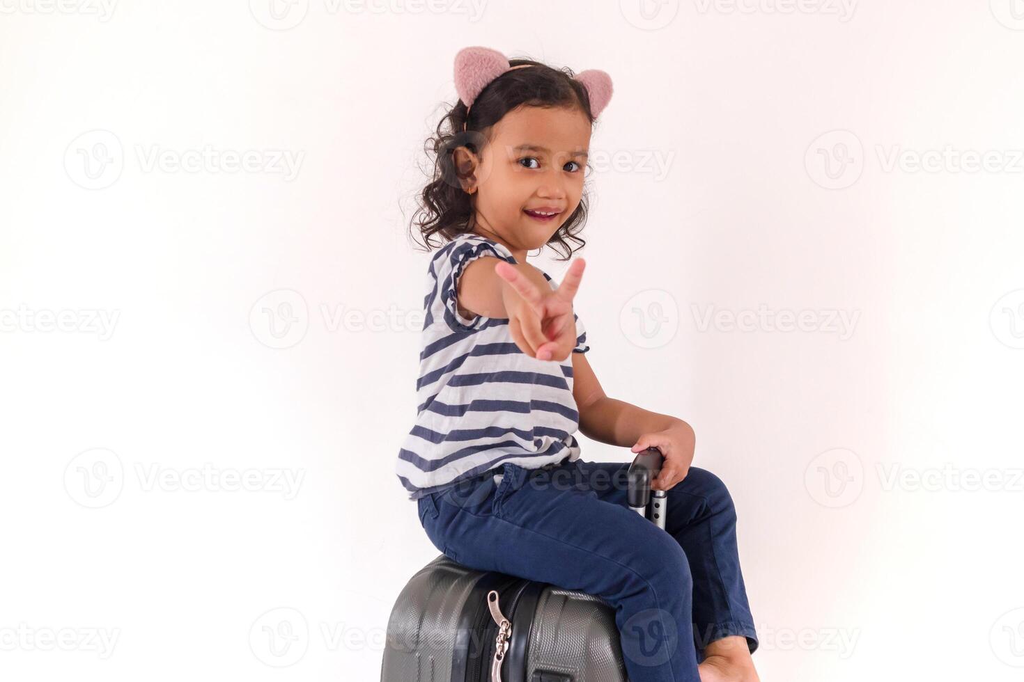 A smiling little Asian girl sitting on a suitcase and showing two fingers or victory sign, on a white background photo