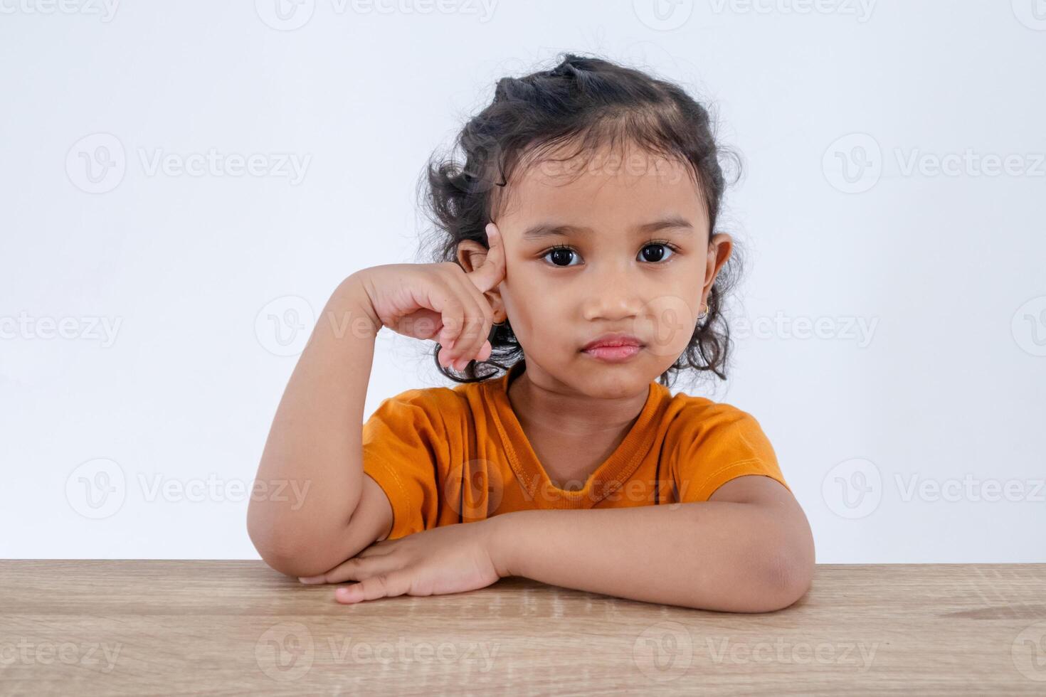 A cute little Asian girl sitting at table and posing with one hand pointing to her head. Confused, thinking, or dreaming concept. photo