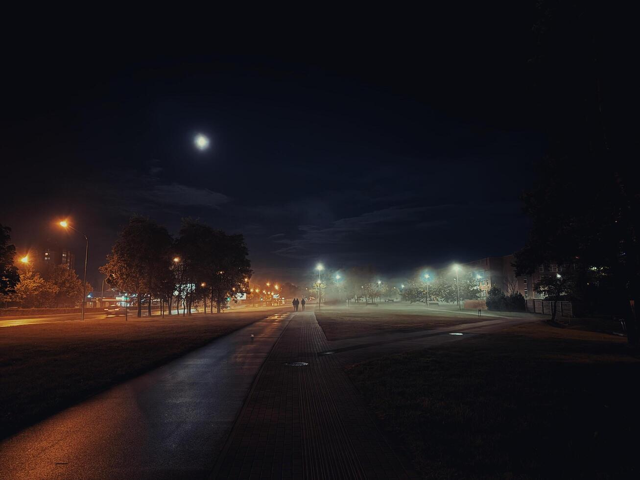 A lone figure walks along a dimly lit path on a foggy night, with the moon shining brightly in the distance. The streetlights cast an ethereal glow on the surrounding trees and grass. photo