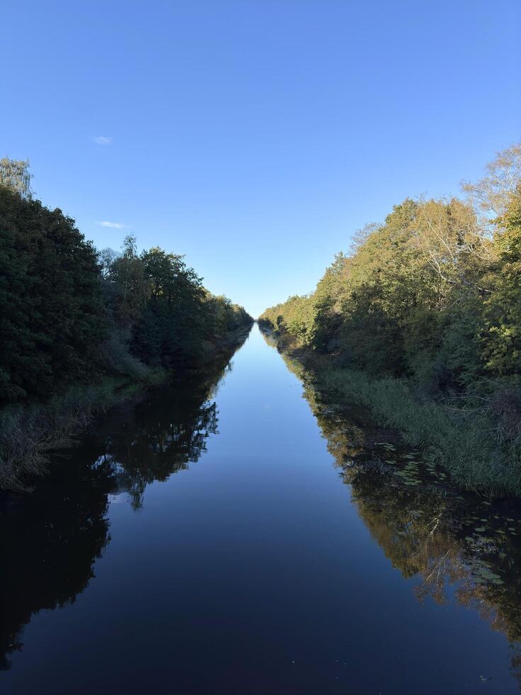 The image shows a calm and still water canal flanked by trees on both sides under a clear blue sky. The reflection of the trees and sky on the water creates a symmetrical visual effect. photo